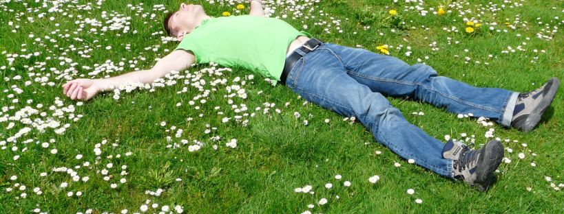 Man laying in green grass with while daisies and some yellow dandelion - he is laying arms and legs apart head back, looking relieved - perhaps experiencing relief while greiving
