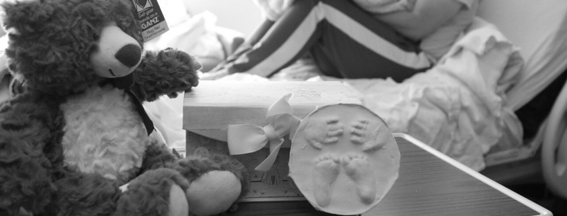 A teddy bear and plaster handprints & footprints on a hospital room tray, dad in the background hospital bed, feeling child loss for the first time