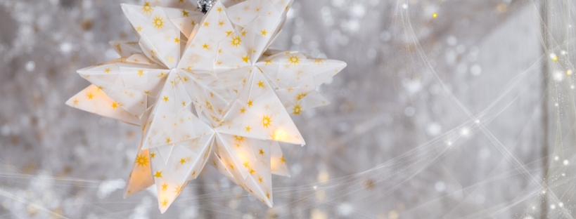 Yellow and white handfolded lantern hung by a sliver pipe cleaners, snowy/spider web type decorations hang behind with tiny lights sparkling, the kind that you might like to share with a child having christmas in hospital.